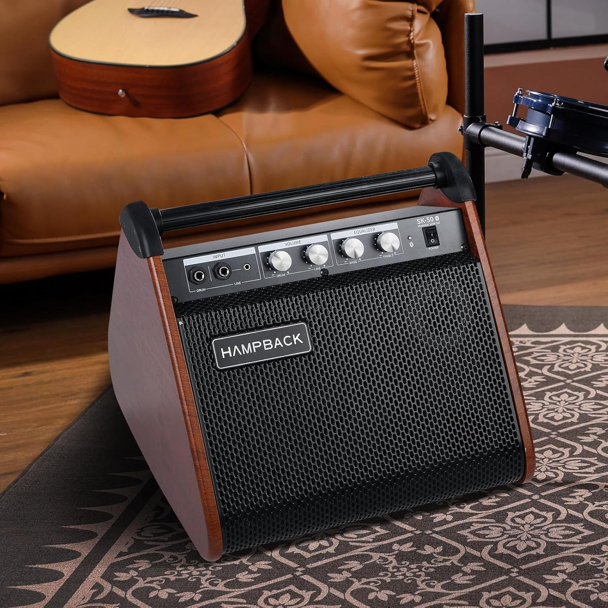 A portable guitar amplifier (Hampback SK-50) with a wood-grain finish and black mesh front grille, placed on a patterned rug in a living room setting with a leather sofa and guitar in the background.